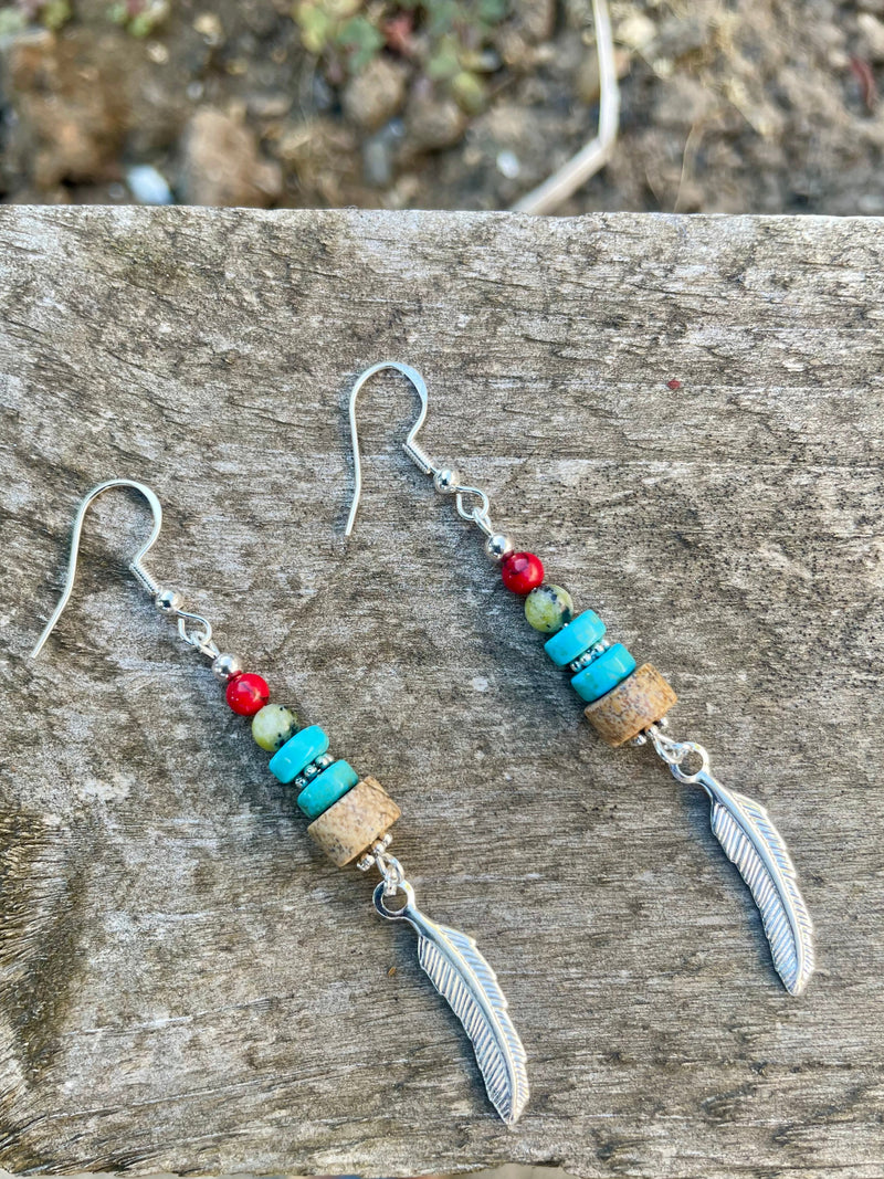 Pair of beaded earrings with feathers on a wooden surface.  Kiona: Earth’s warmth and water’s serenity captured in beads, crowned with a silver feather charm.