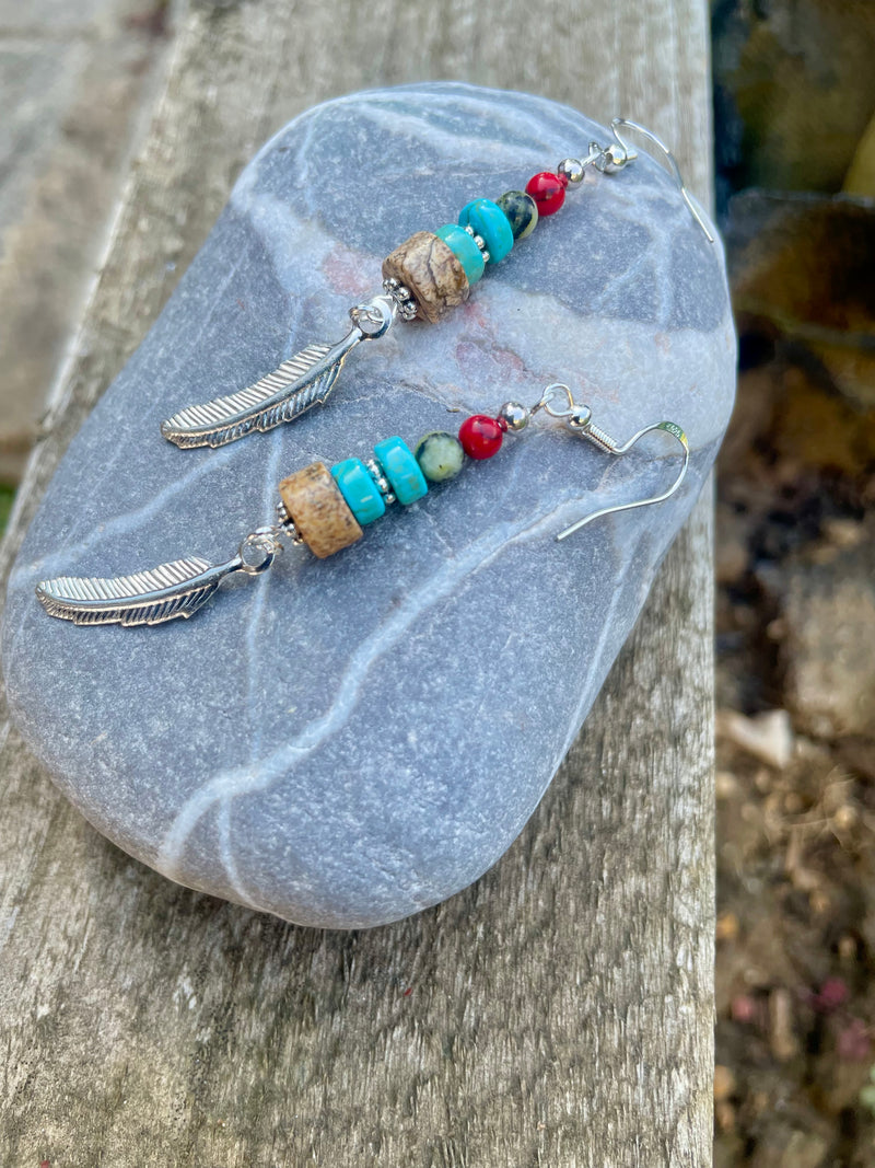 Beaded earrings with feathers on a stone surface.  Kiona: Earth’s warmth and water’s serenity captured in beads, crowned with a silver feather charm.
