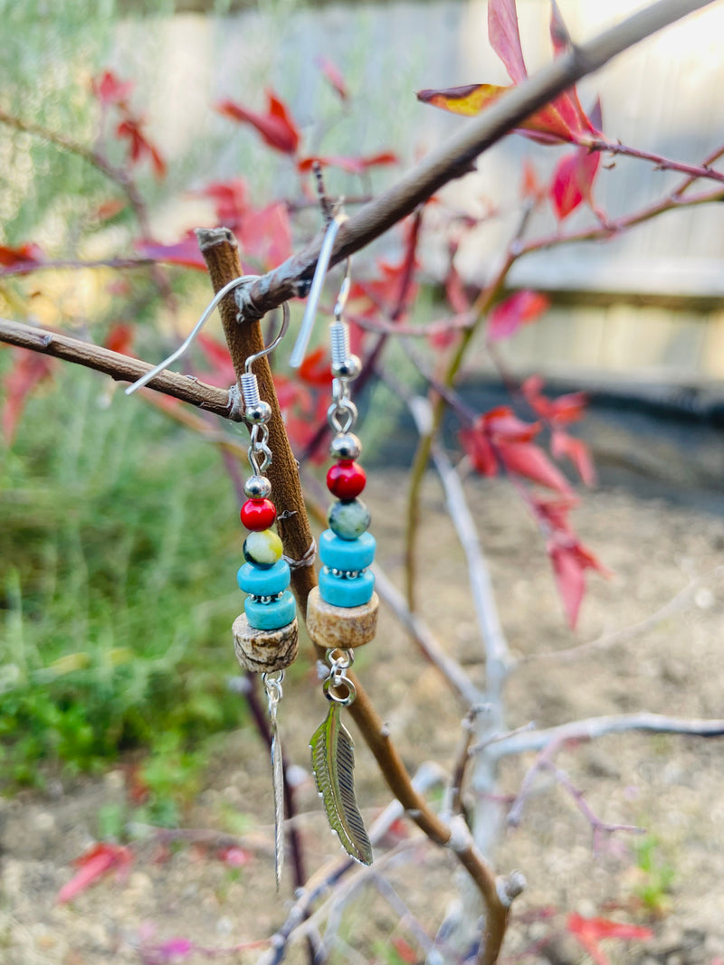 Beaded earrings with a feather charm hanging on a branch with leaves.  Kiona: Earth’s warmth and water’s serenity captured in beads, crowned with a silver feather charm.